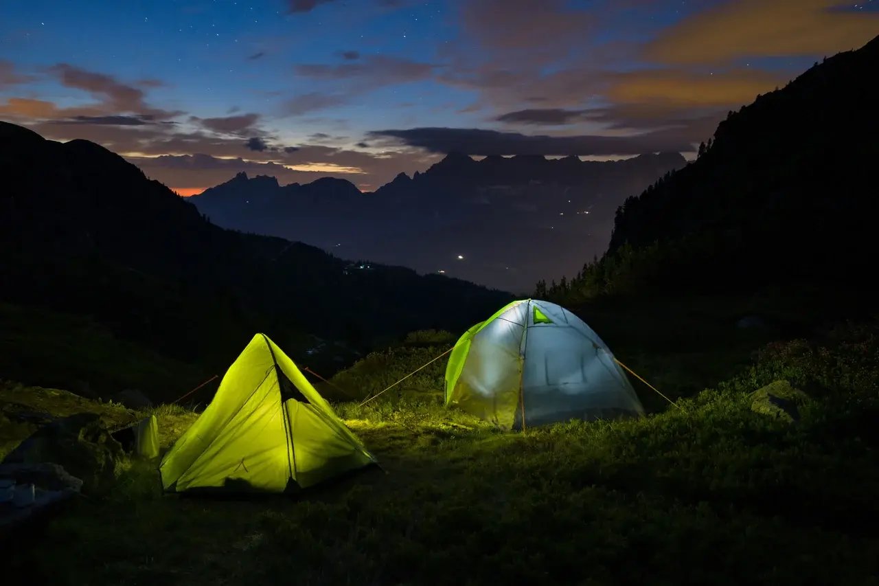Two brightly lit tents are set up in a mountainous landscape at dusk, with a starry sky and distant peaks visible in the background.