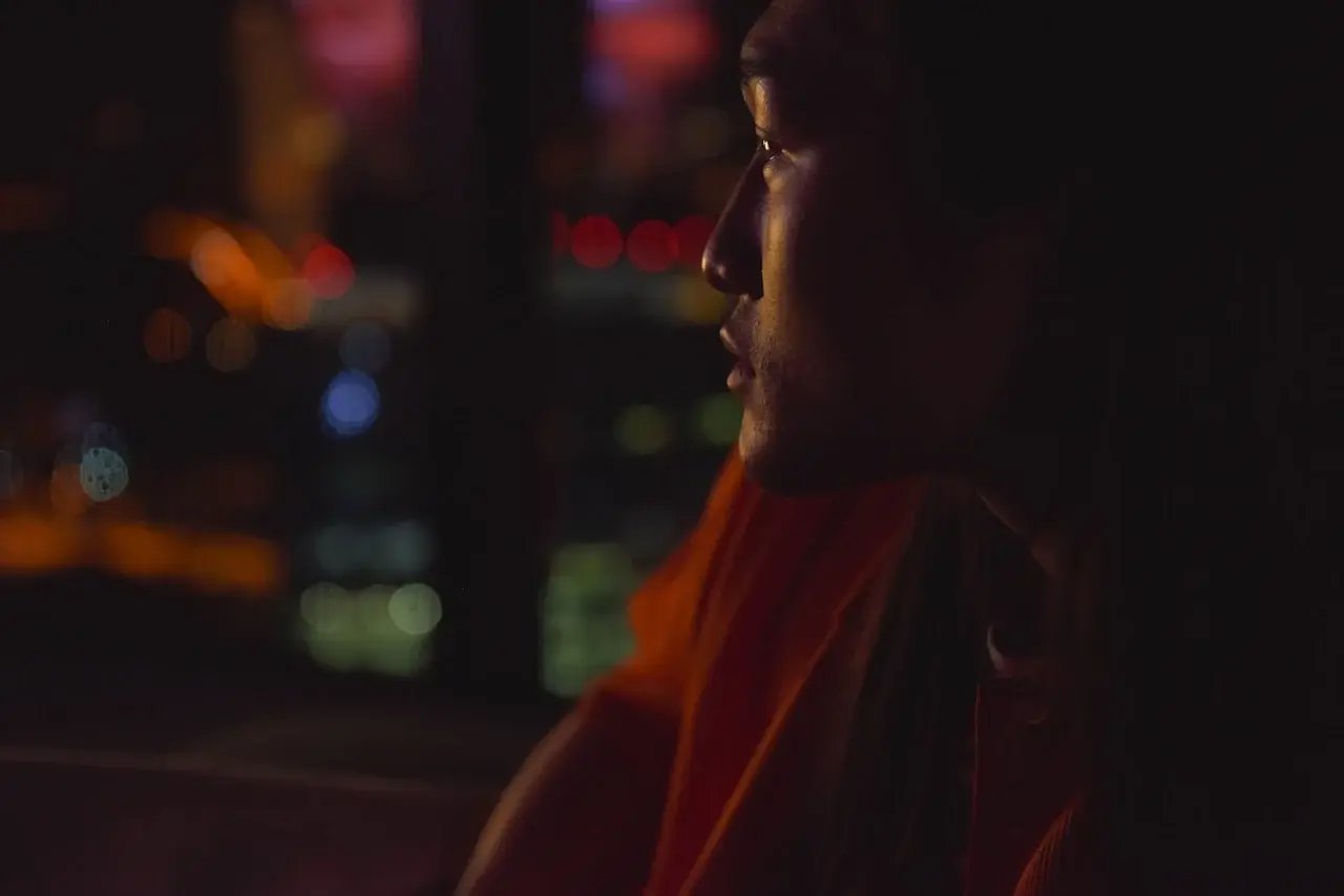 Side-profile portrait of a person in soft, warm lighting, wearing an orange shirt and gazing out toward a blurred nighttime cityscape with colorful bokeh lights in red, orange, blue, and green.