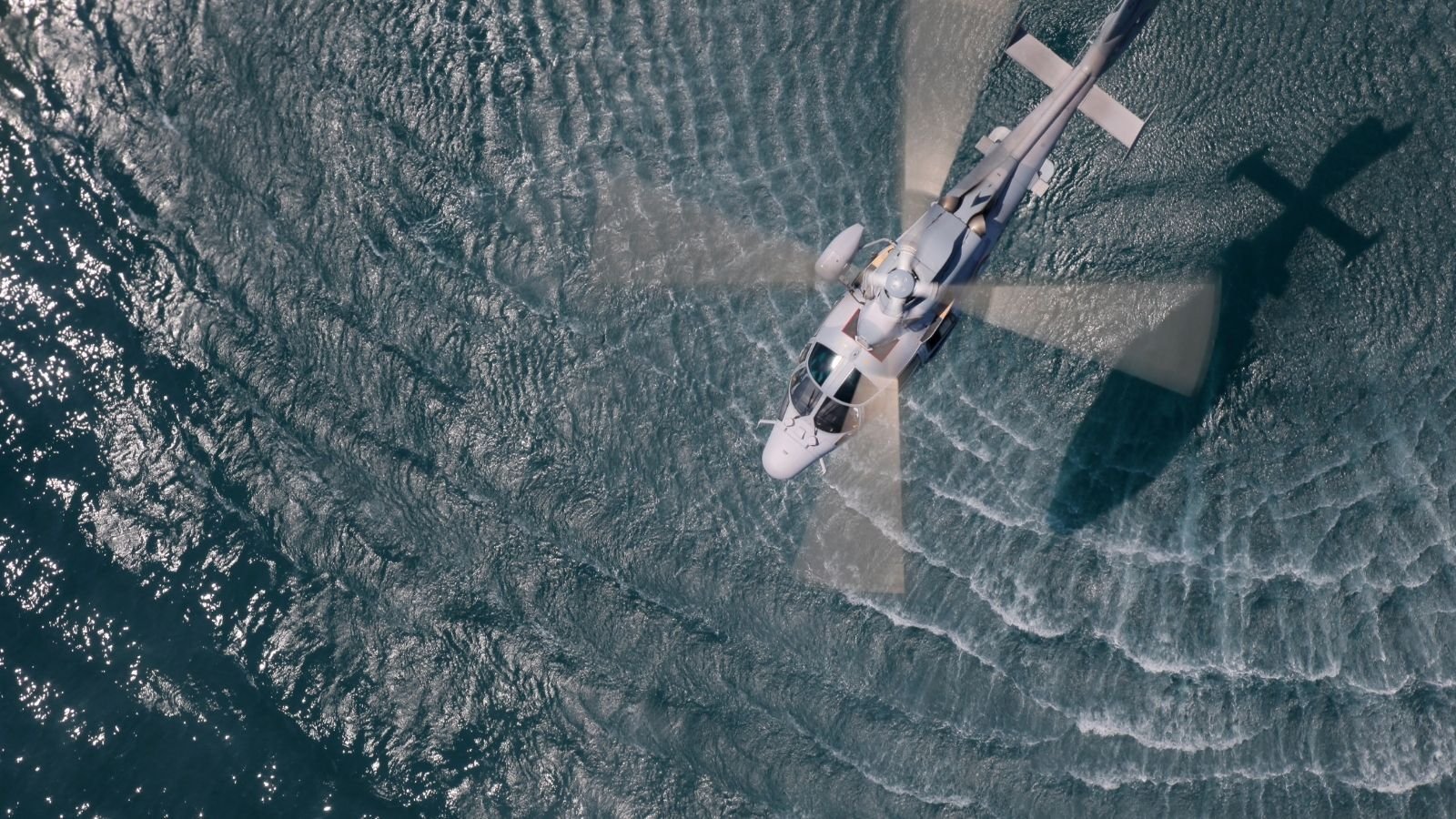 A helicopter is flying over the ocean, creating rippling water patterns below. The bright sun reflects off the water, casting the helicopter's shadow.