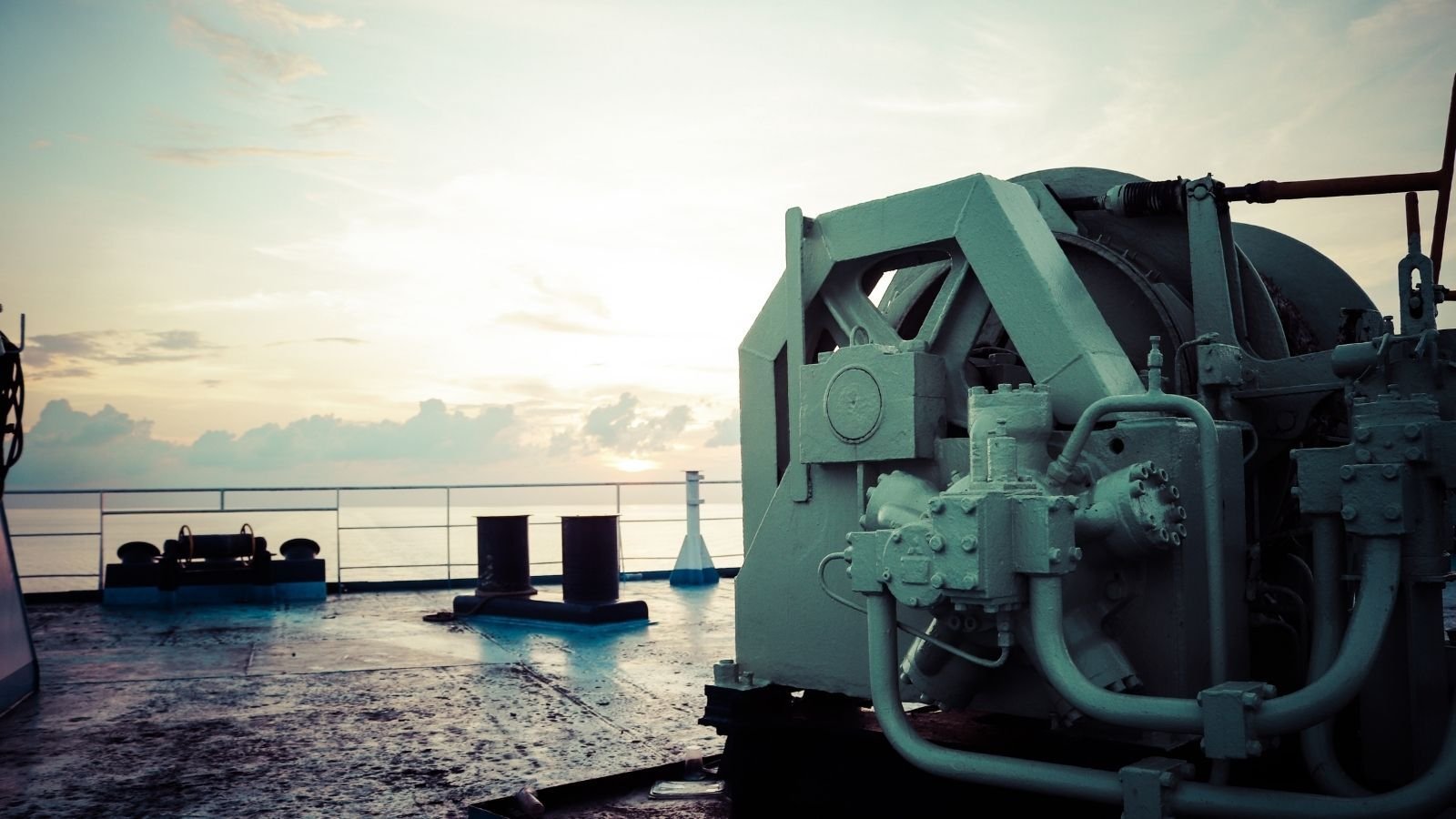 Deck machinery on a ship at sunrise, featuring large winches and equipment. The sky is pastel with soft clouds.
