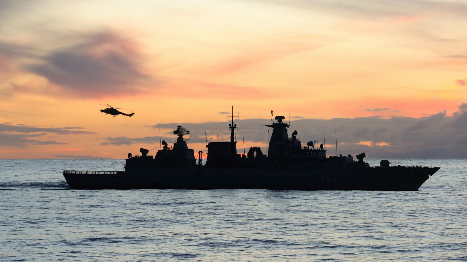 Silhouette of a naval warship sailing at sea with a helicopter flying above against a colorful sunset sky.