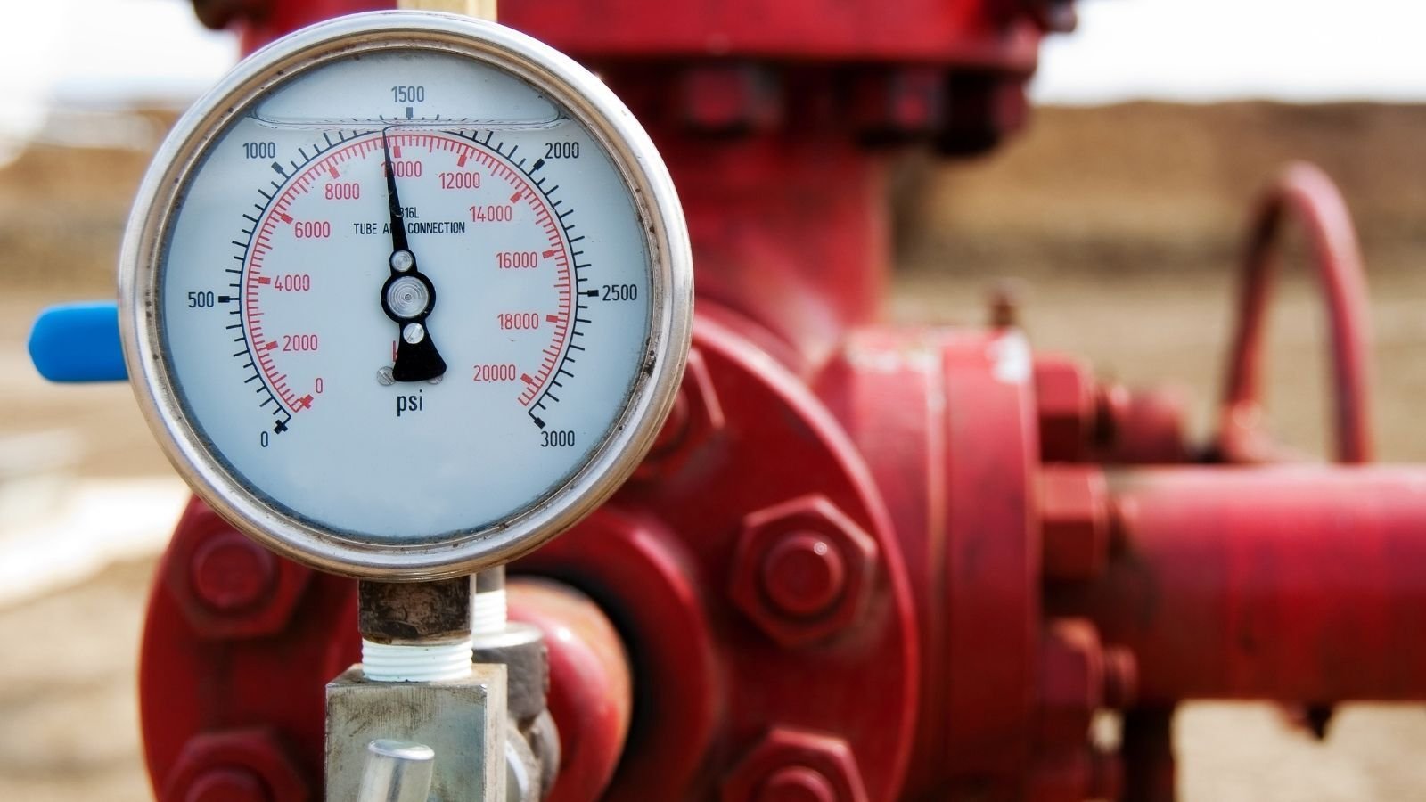 Close-up of a pressure gauge reading in psi, mounted on a large red industrial pipe system, set against a blurred outdoor background. The gauge needle points to around 1500 psi.