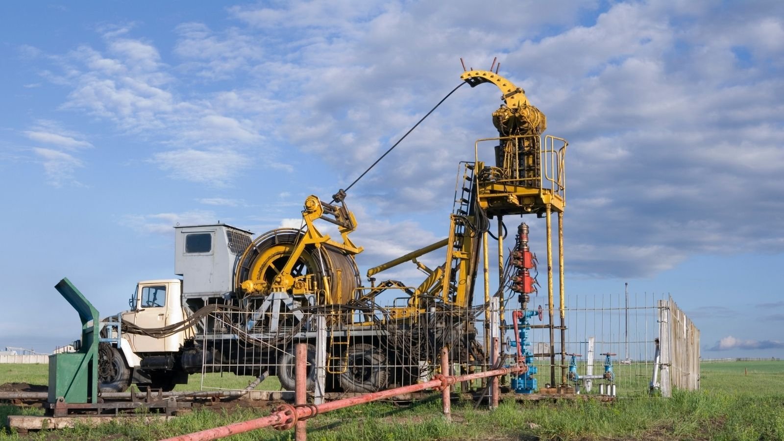 A yellow oil rig with a truck is set in a grassy field under a partly cloudy sky. The machinery includes large wheels and cables.