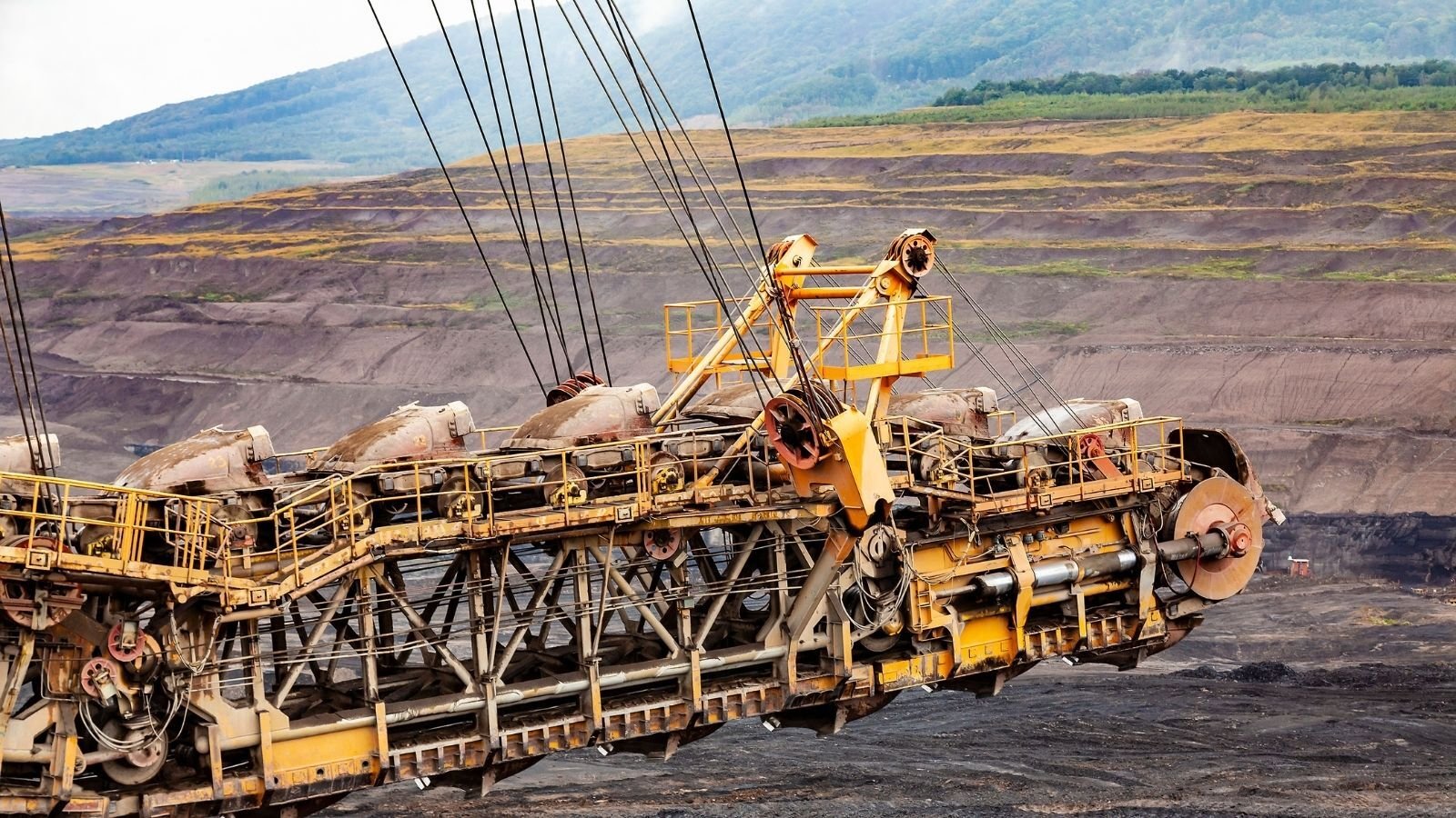 A massive industrial excavator operates in an open-pit mine. The machinery is detailed and imposing, set against layered earth and distant green hills.