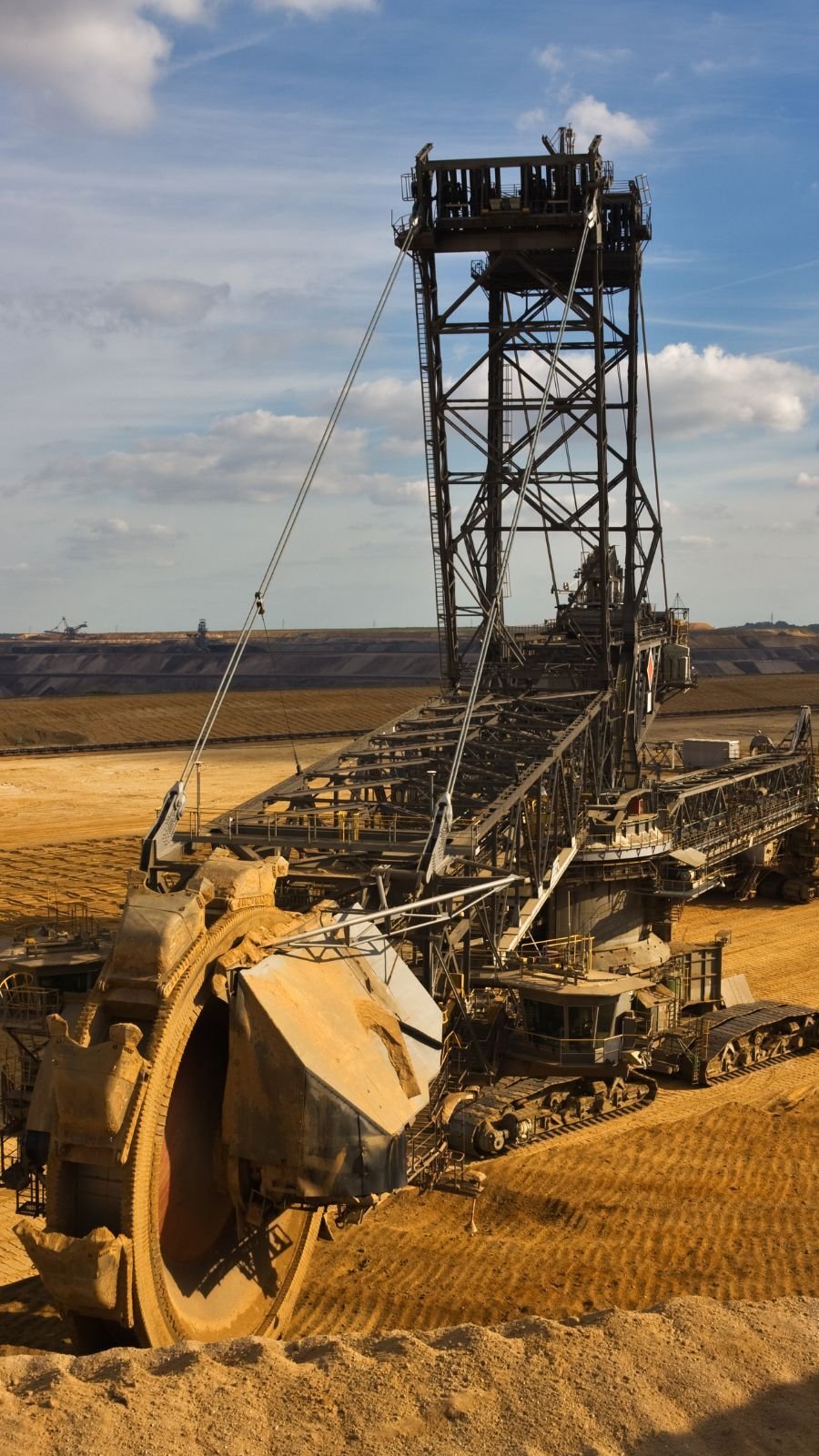 Massive excavator with large rotating bucket-wheel on dirt landscape, set against a bright blue sky with clouds.