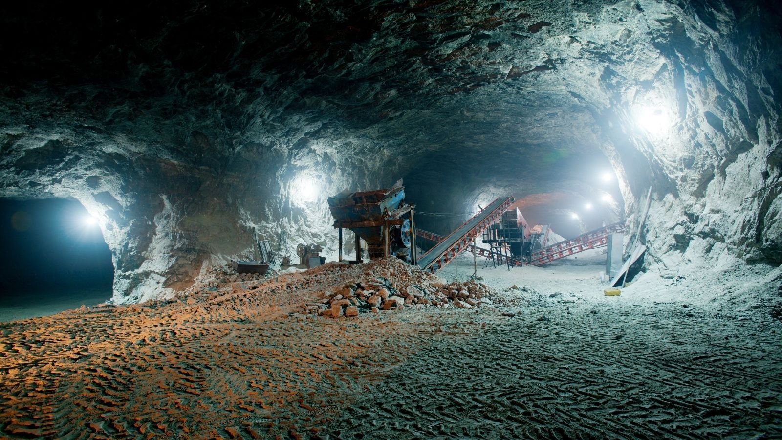 Underground mine with rugged rock walls, illuminated by bright lights. Conveyors and machinery are visible.