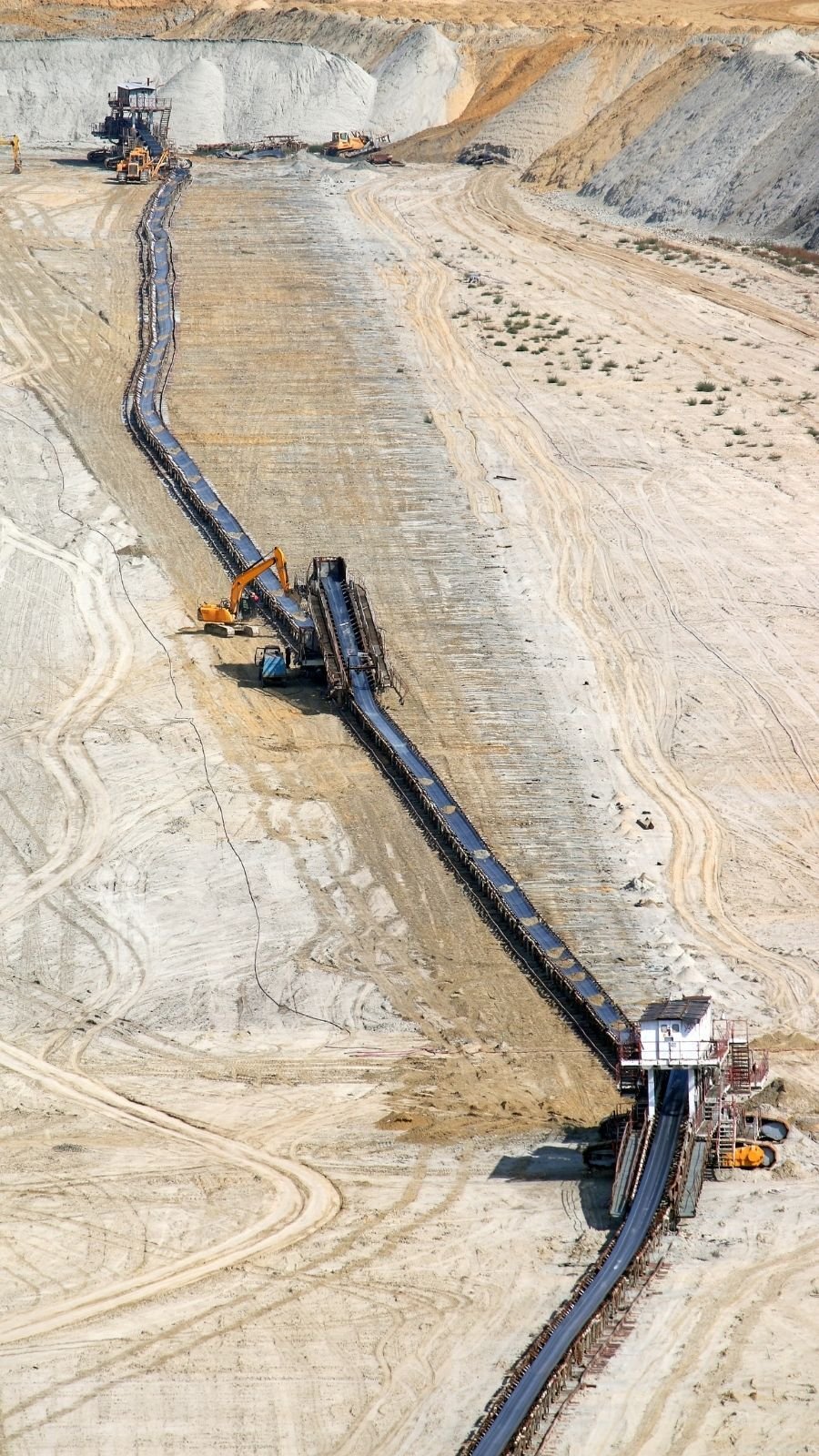 Aerial view of a large mining site with a long conveyor belt stretching across sandy terrain. Excavators work nearby under a clear sky.