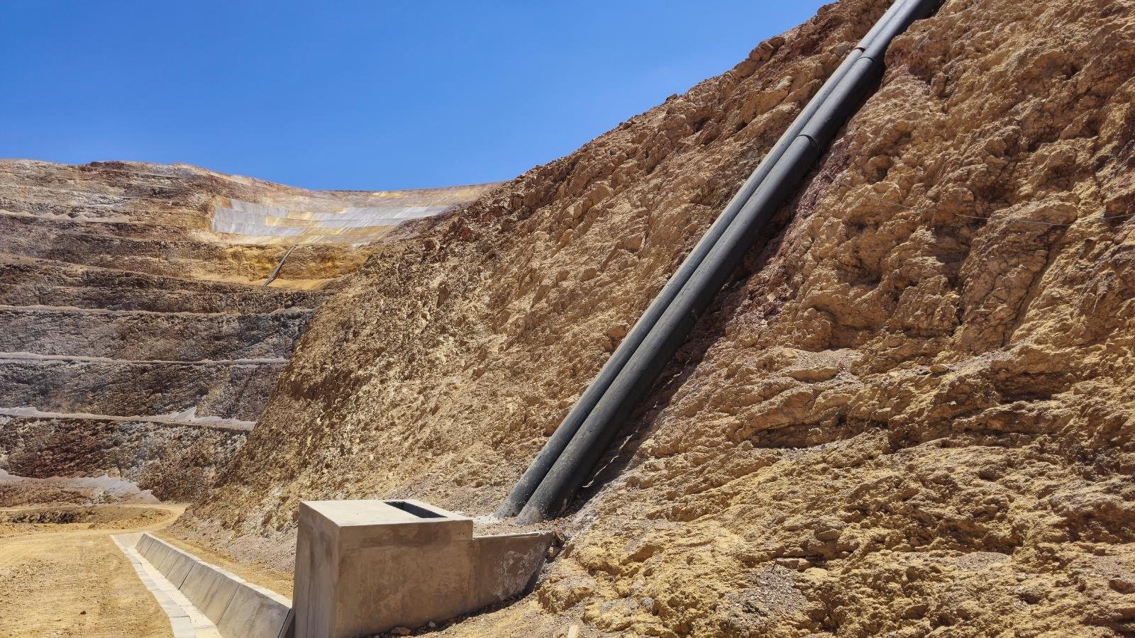 A rugged, rocky landscape with a steep incline showing large black pipes secured to it.