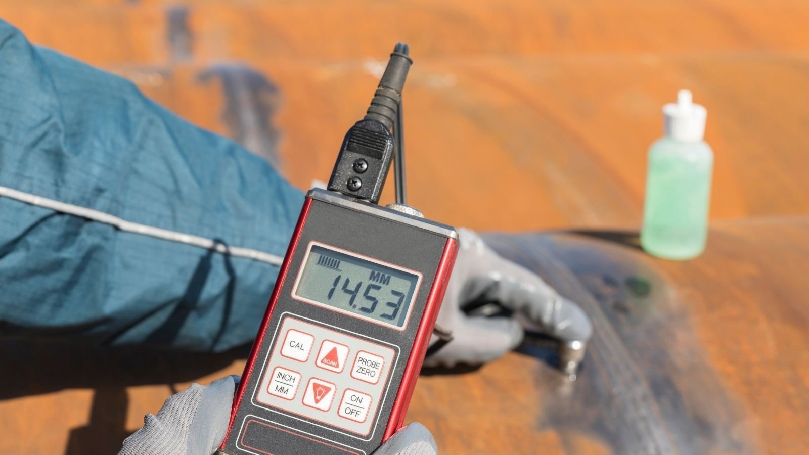 A person in gloves uses a digital gauge to measure thickness, displaying 145.3 mm, on rust-colored metal. A bottle with liquid is in the background.