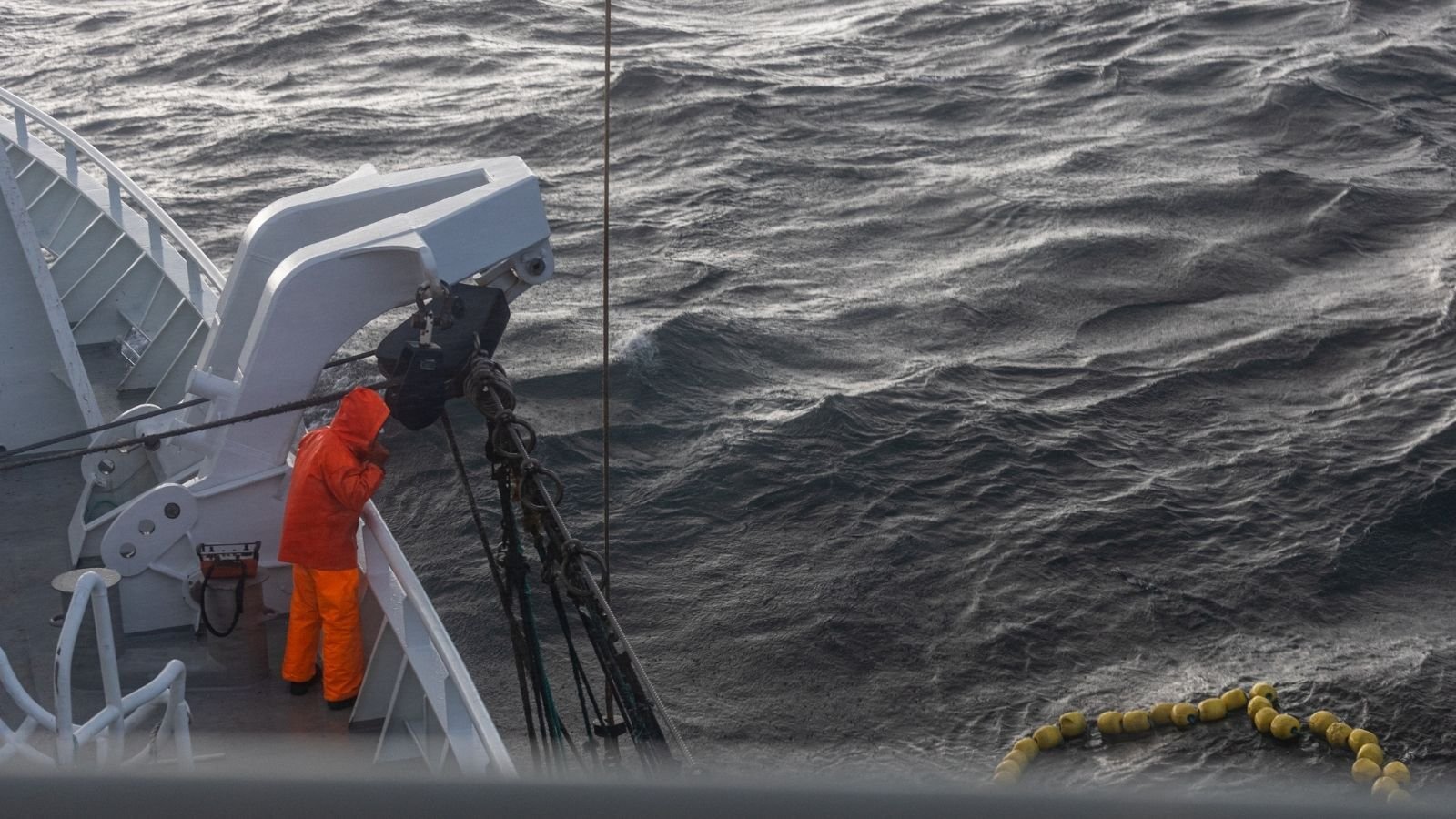 A person in bright orange rain gear stands on a ship's deck, facing choppy, dark ocean waves. A buoy line is visible on the water, creating a tense, isolated mood.