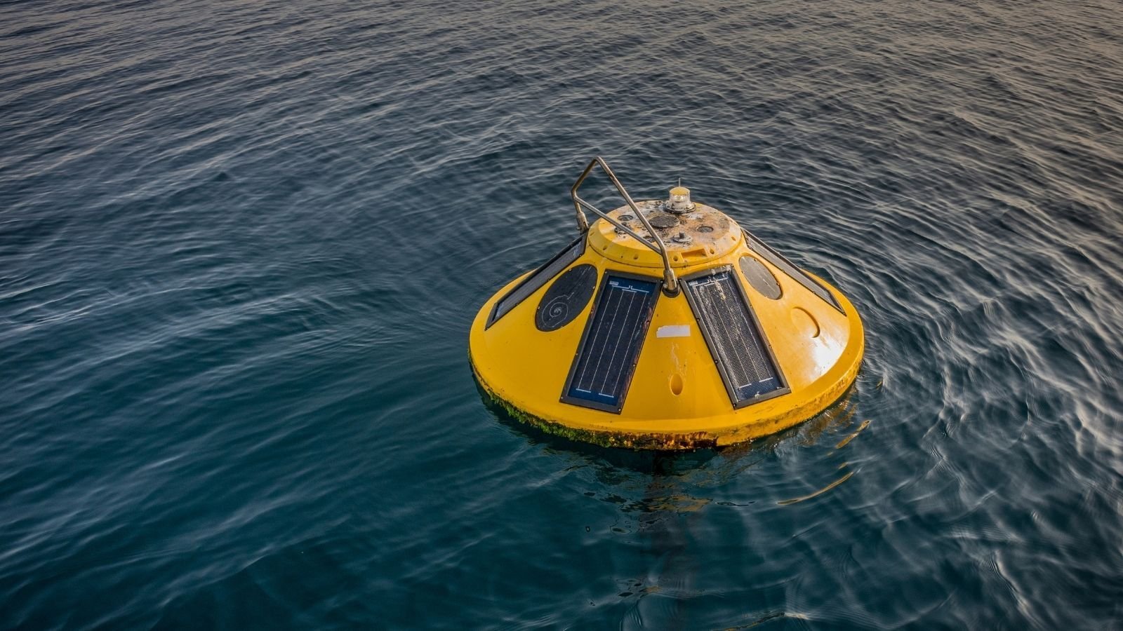 A yellow buoy with solar panels floats on calm blue water, creating a serene and peaceful atmosphere. The sunlight reflects gently off the water's surface.