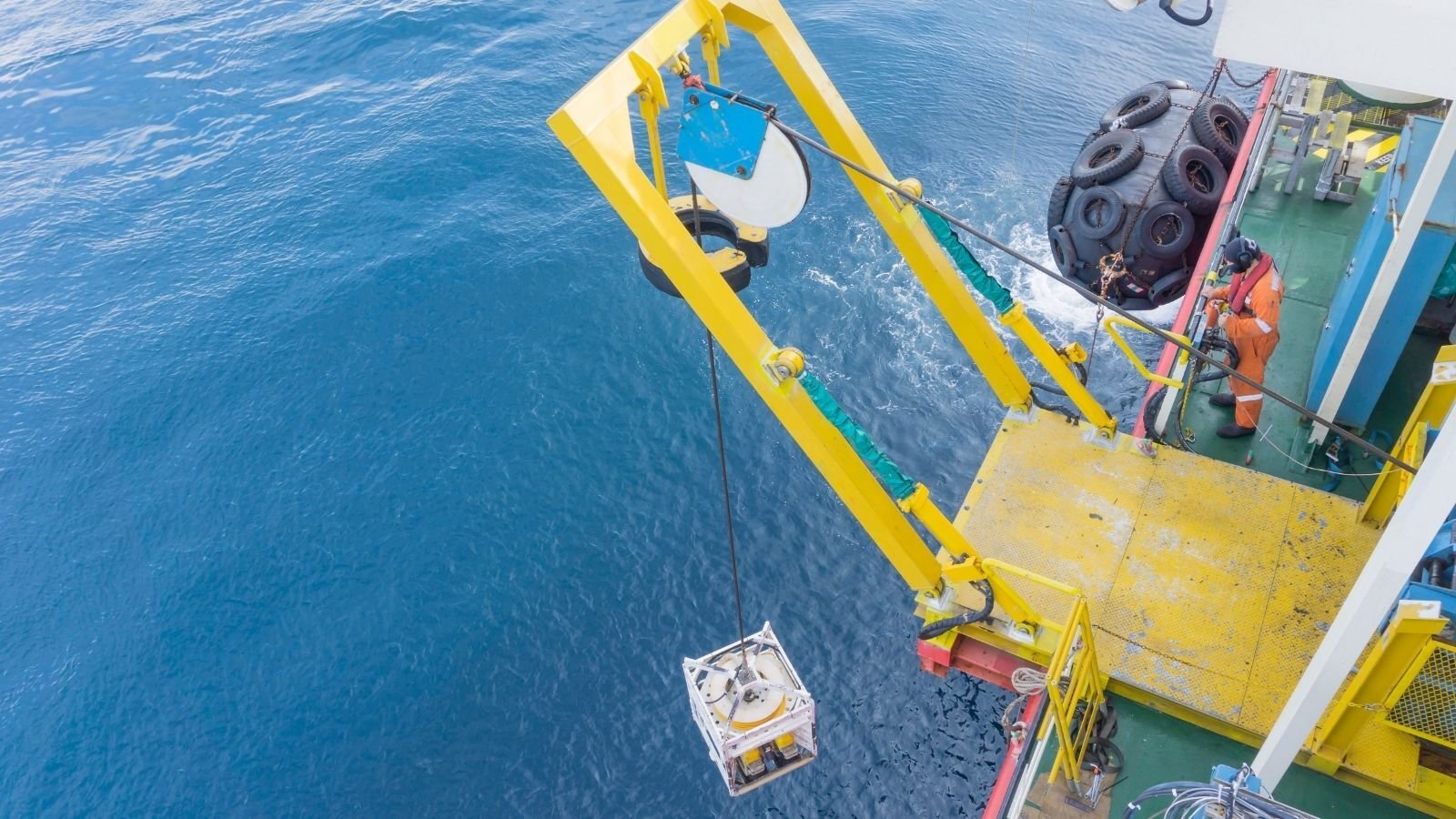 An overhead view of a yellow crane on a ship, lowering equipment into the deep blue sea. A worker in orange coveralls oversees the operation.