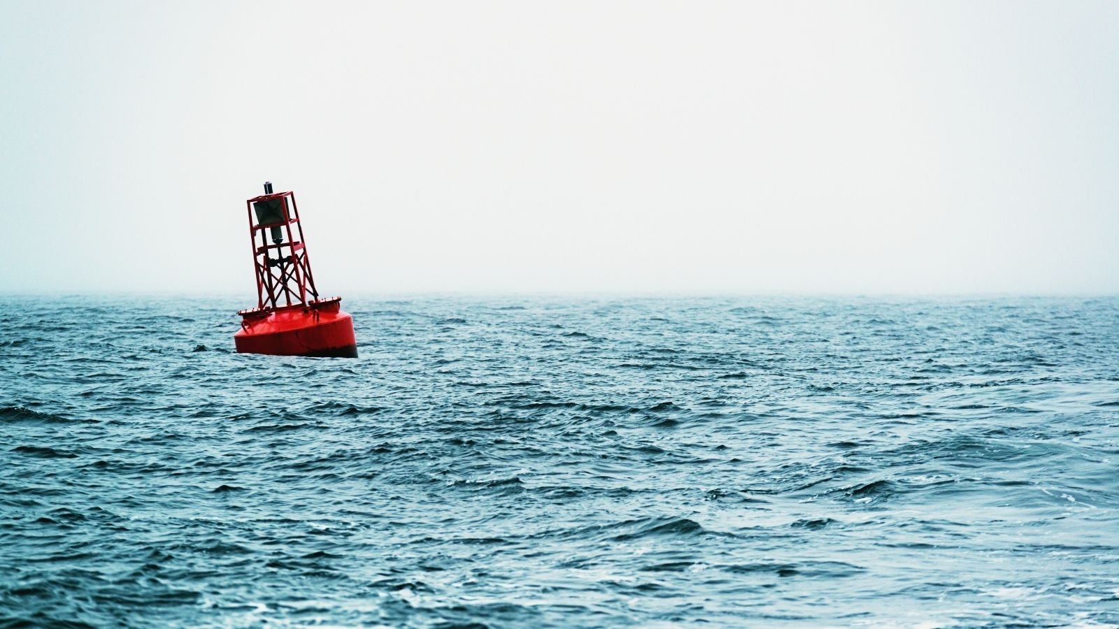 A solitary red buoy gently tilts amidst vast, choppy ocean waves under a light gray sky.