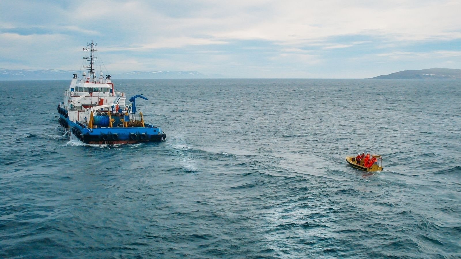 A large blue and white ship sails in the open sea, observed by a small yellow lifeboat with people in red life jackets, under a cloudy sky.