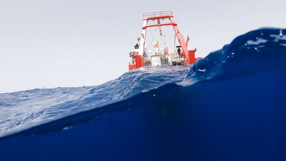 A vibrant red ship with industrial equipment is seen from a low angle, partially submerged amid deep blue ocean waves, set against a clear sky.