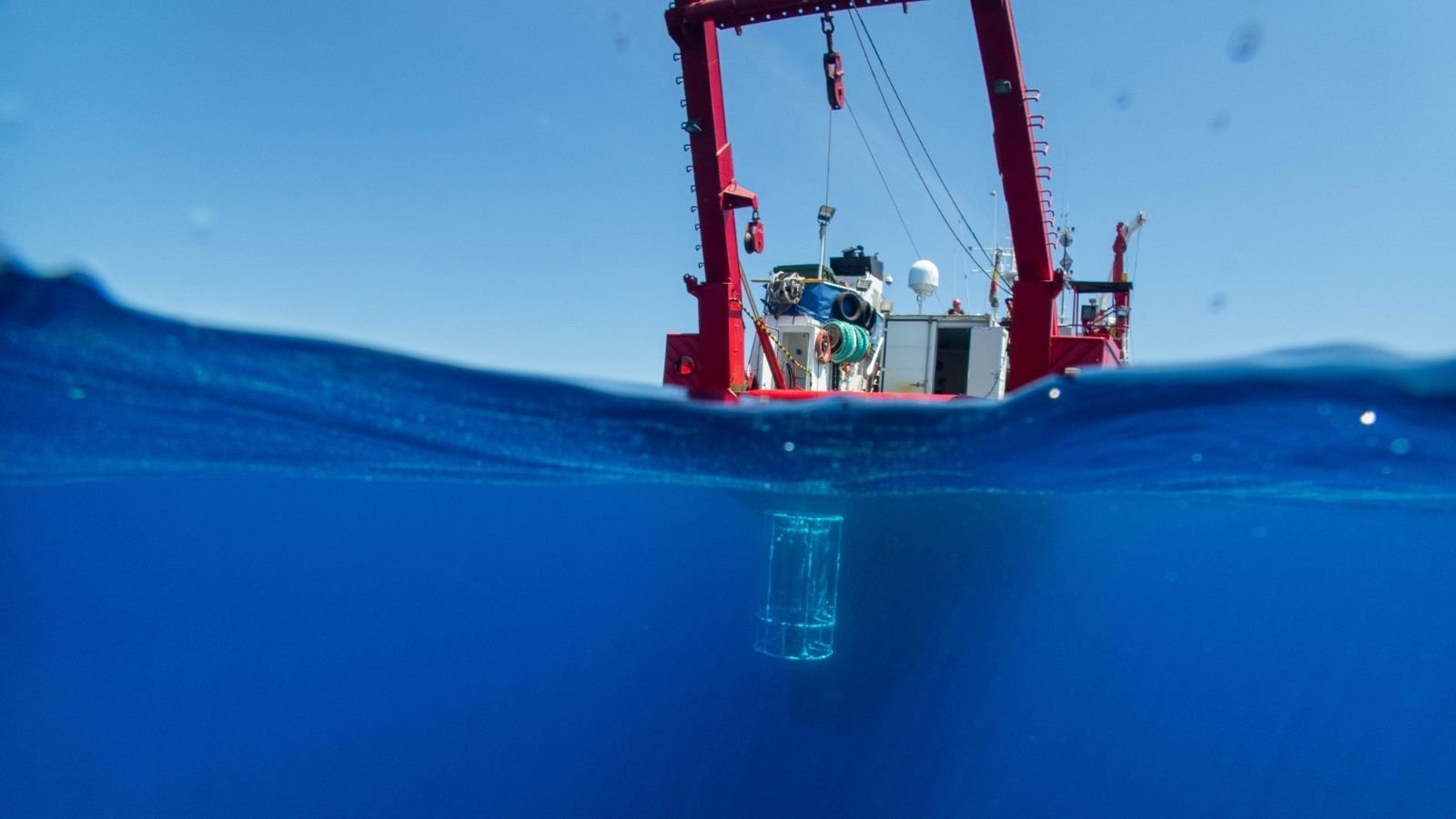 A red research vessel on the ocean with a calm sea. Beneath the surface, a transparent container is partially submerged, indicating marine exploration.