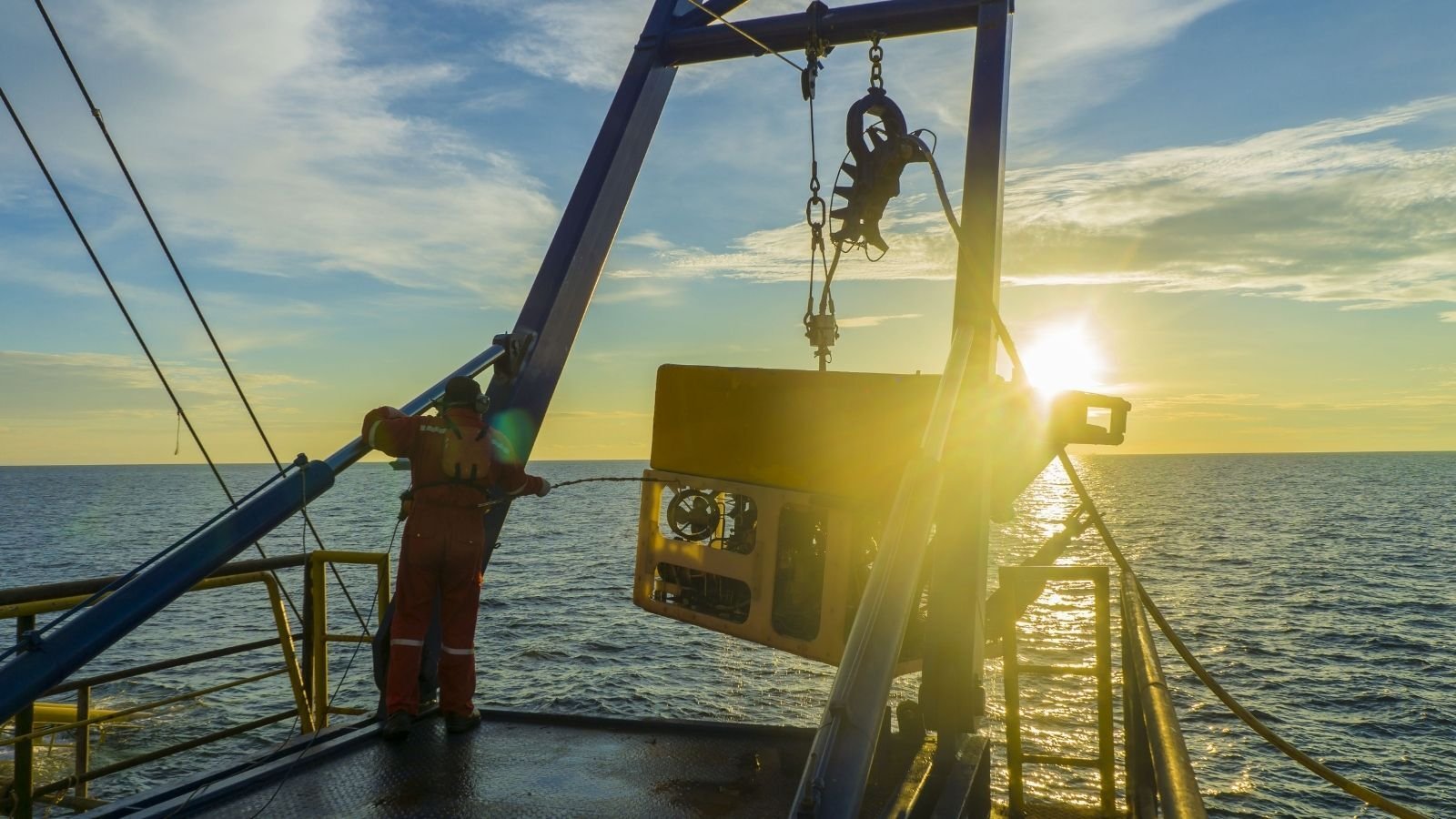 A person in an orange suit stands on a boat, overseeing a crane lowering equipment into the sea. The sun sets on the horizon, casting a warm glow.