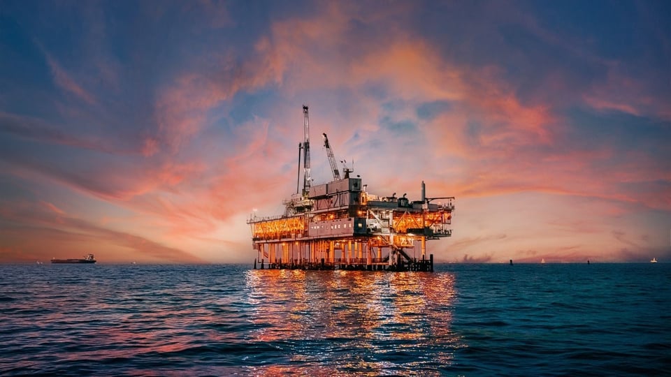An illuminated offshore oil rig stands against a vibrant sunset sky, reflecting on calm ocean waters.