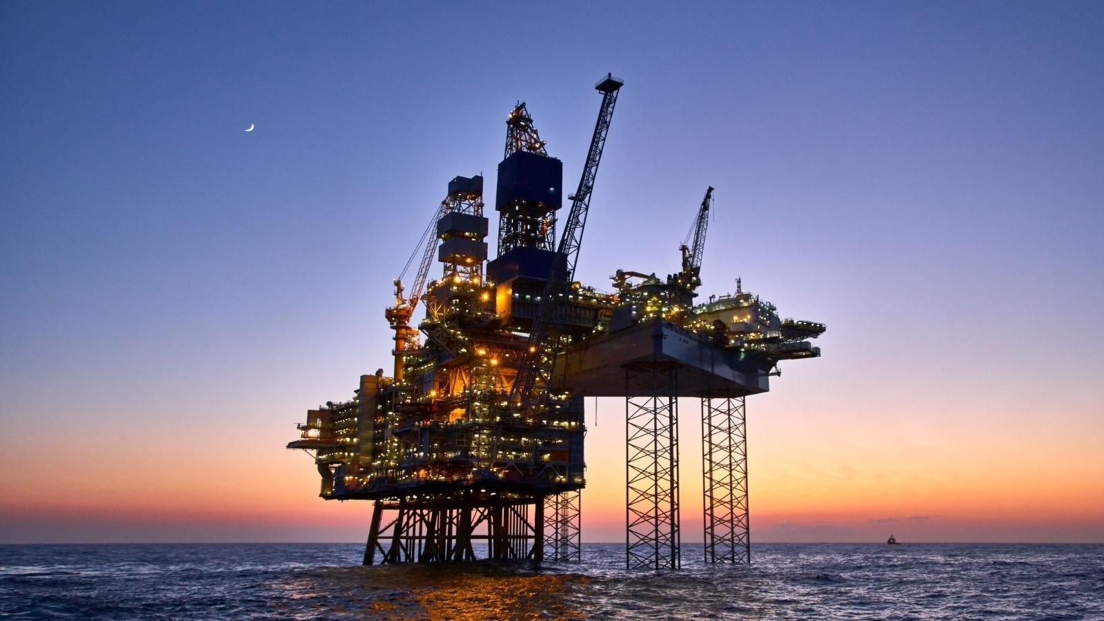 An offshore oil rig illuminated against a twilight sky, with a crescent moon visible.