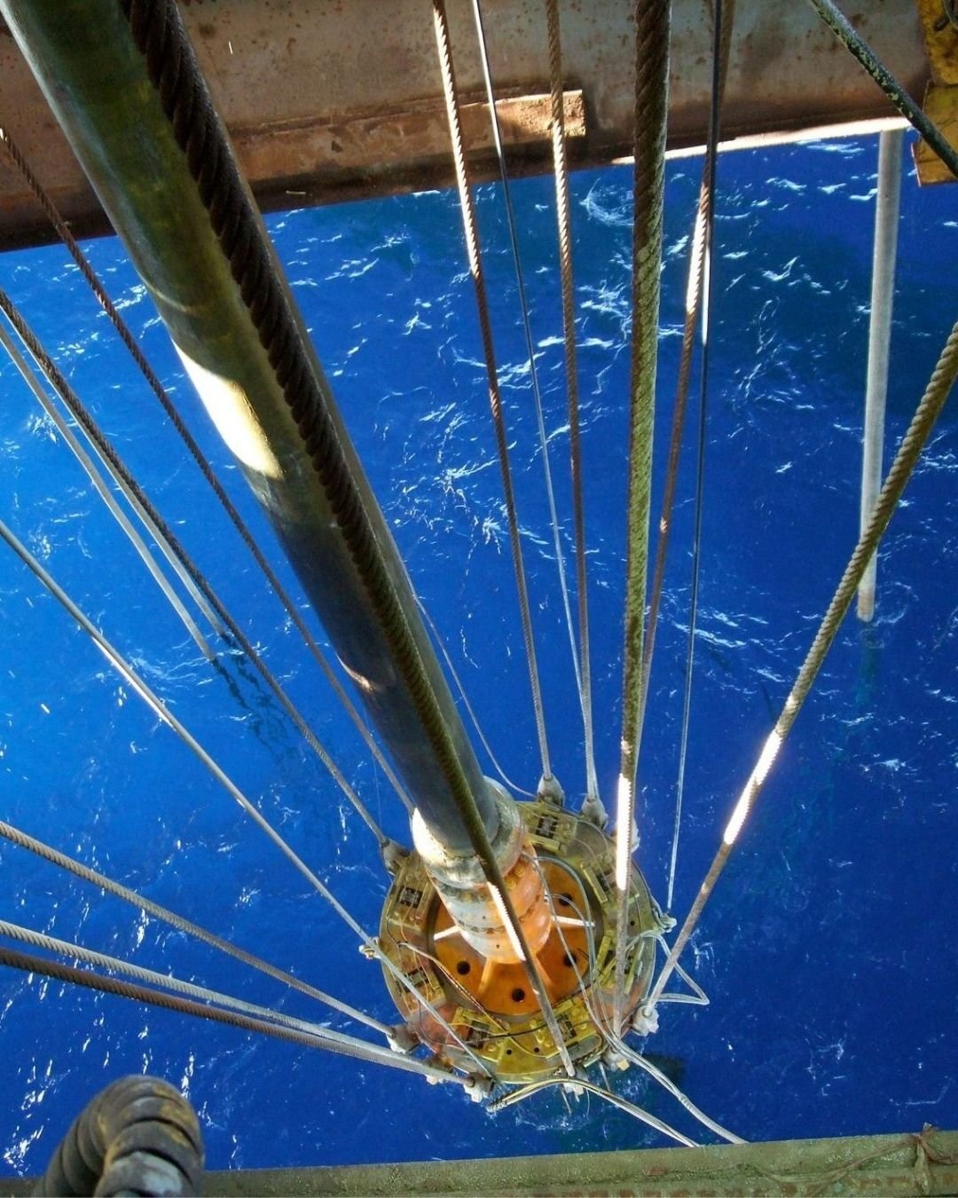 Looking down from an offshore oil rig, cables and pipes extend towards the vibrant blue ocean below, conveying industrial scale and maritime context.