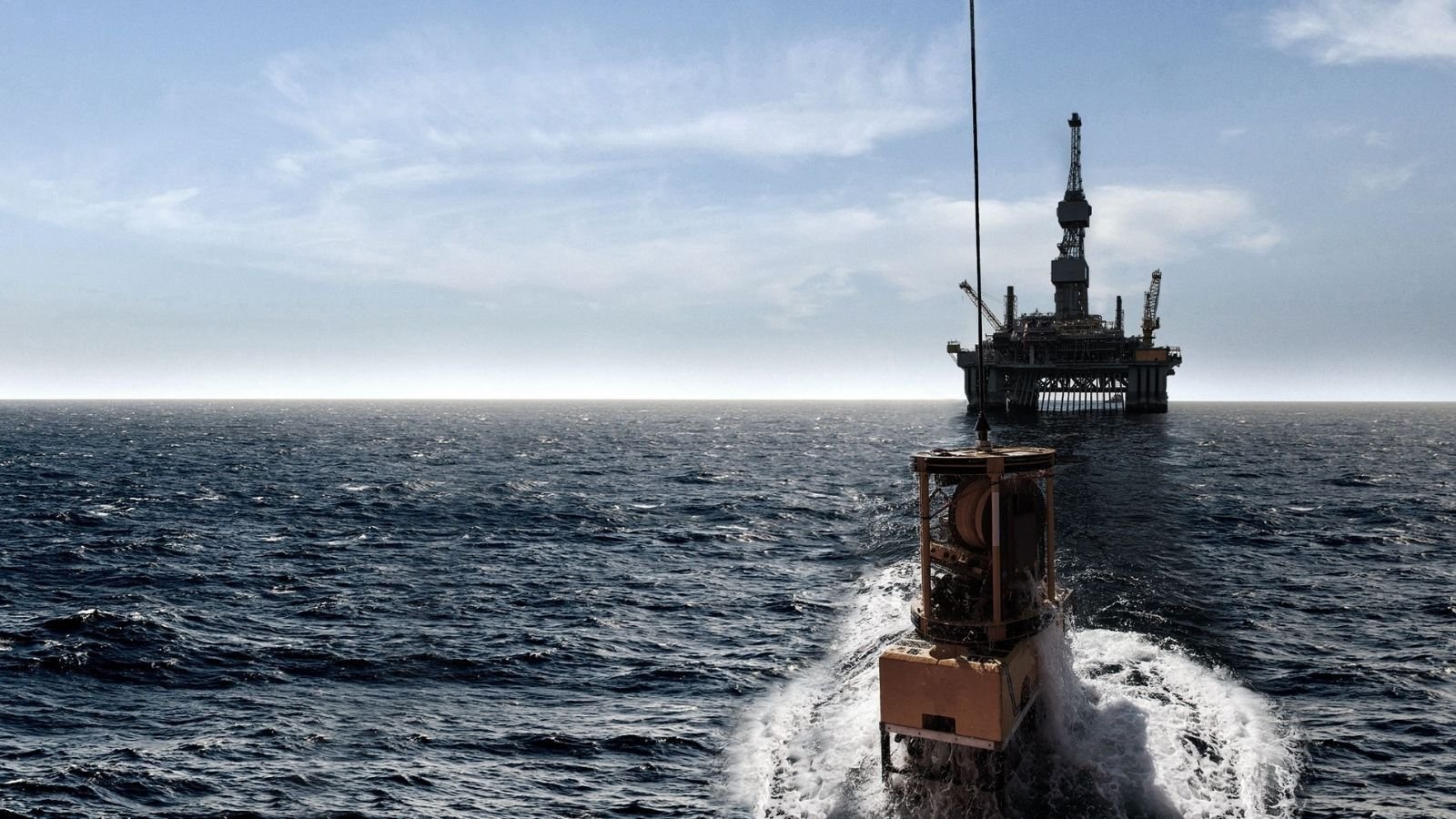 A boat moves through ocean waves towards an offshore oil rig, with a clear sky and distant horizon in the background.