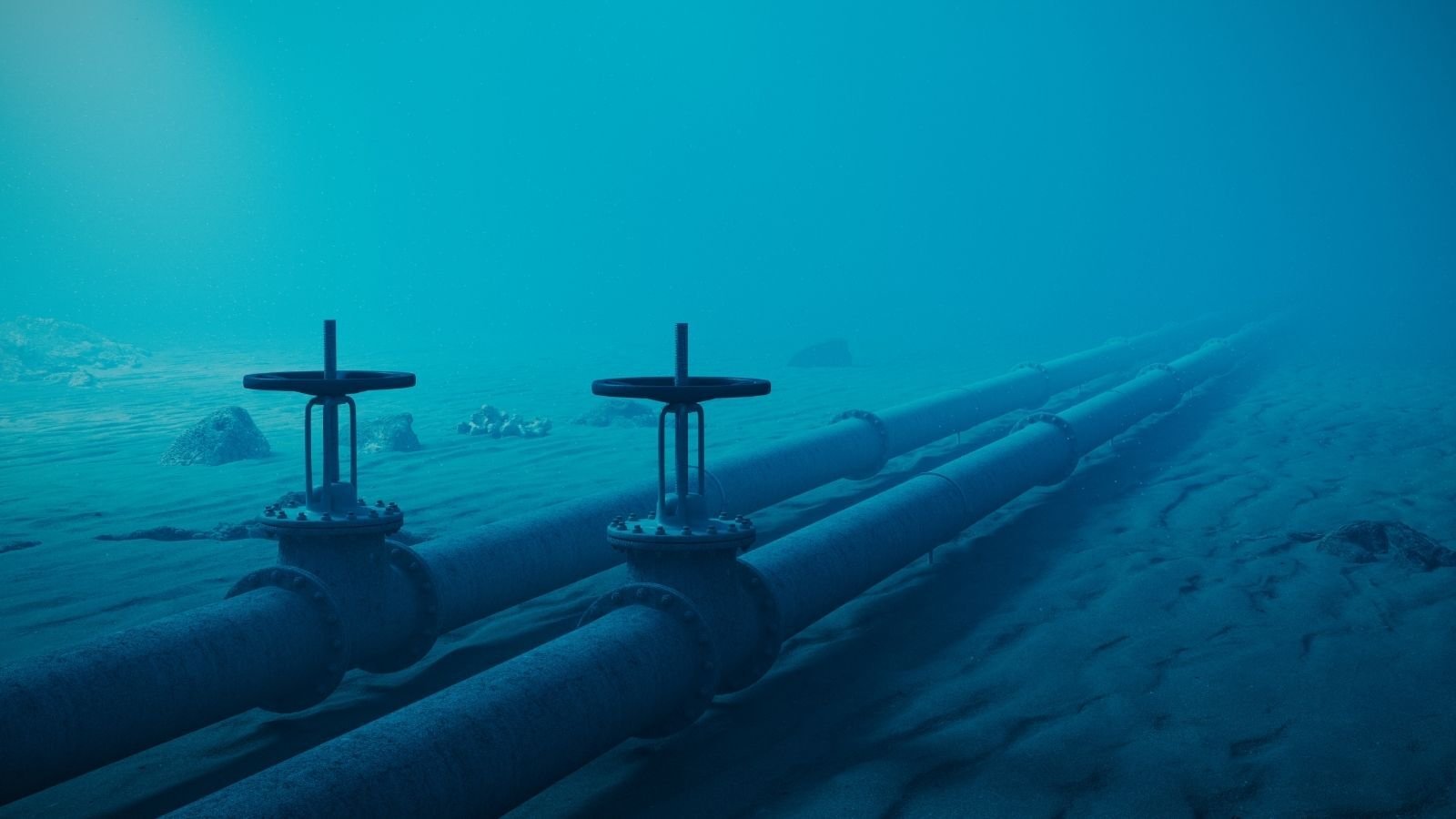 Underwater image showing two parallel pipelines with large valve wheels resting on a sandy seabed.