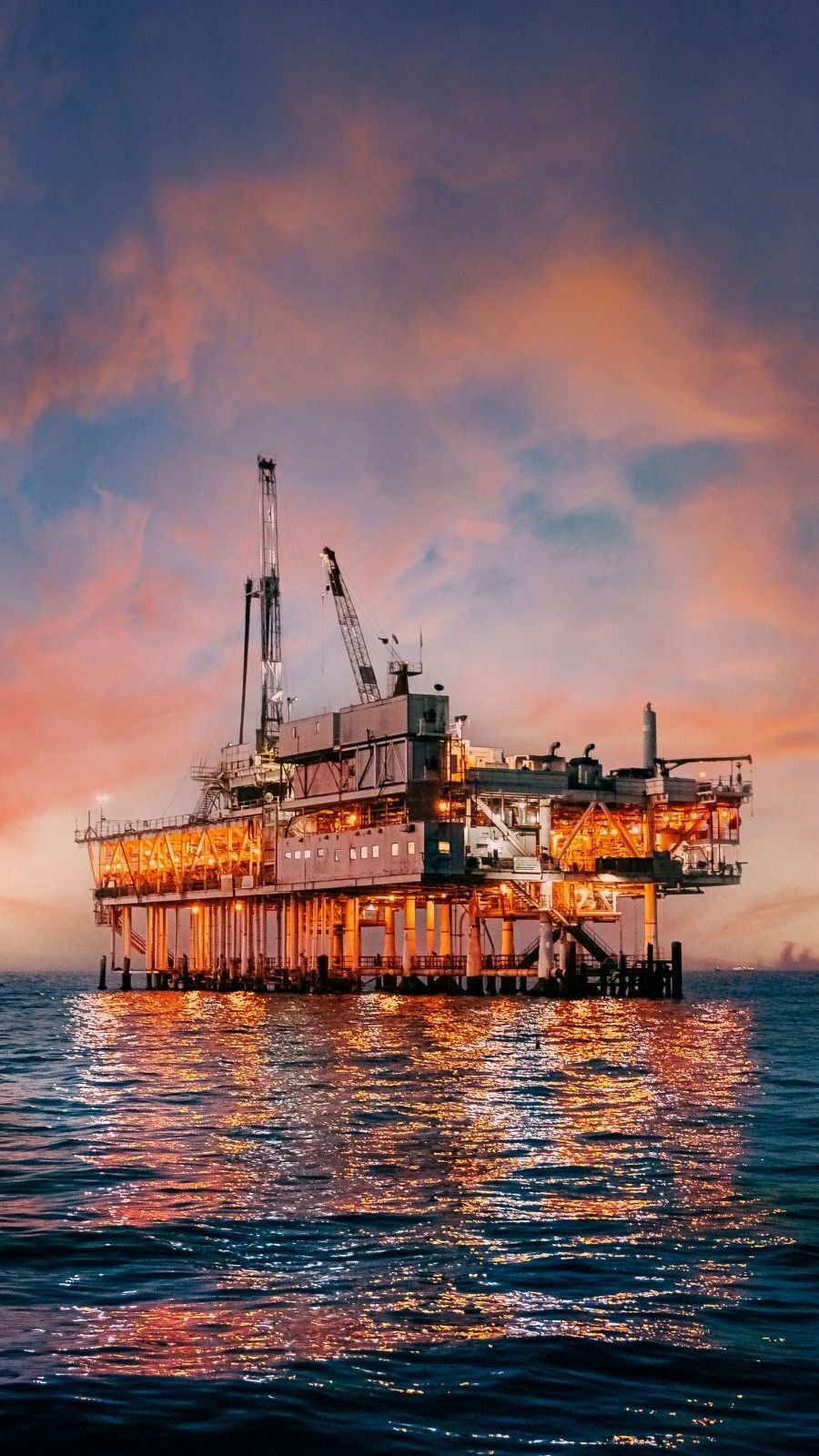 Offshore oil rig illuminated against a vibrant sunset sky, reflecting on the calm ocean.