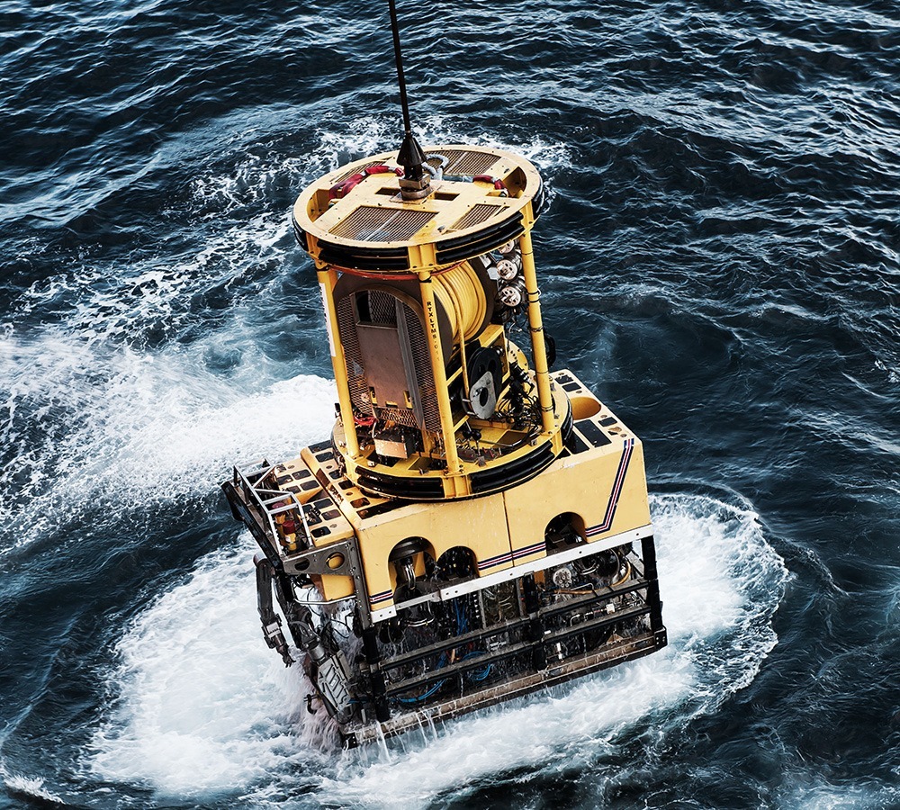 A large, yellow, cylindrical marine structure is partially submerged in the choppy ocean, creating splashes around it. The scene conveys industrial activity.