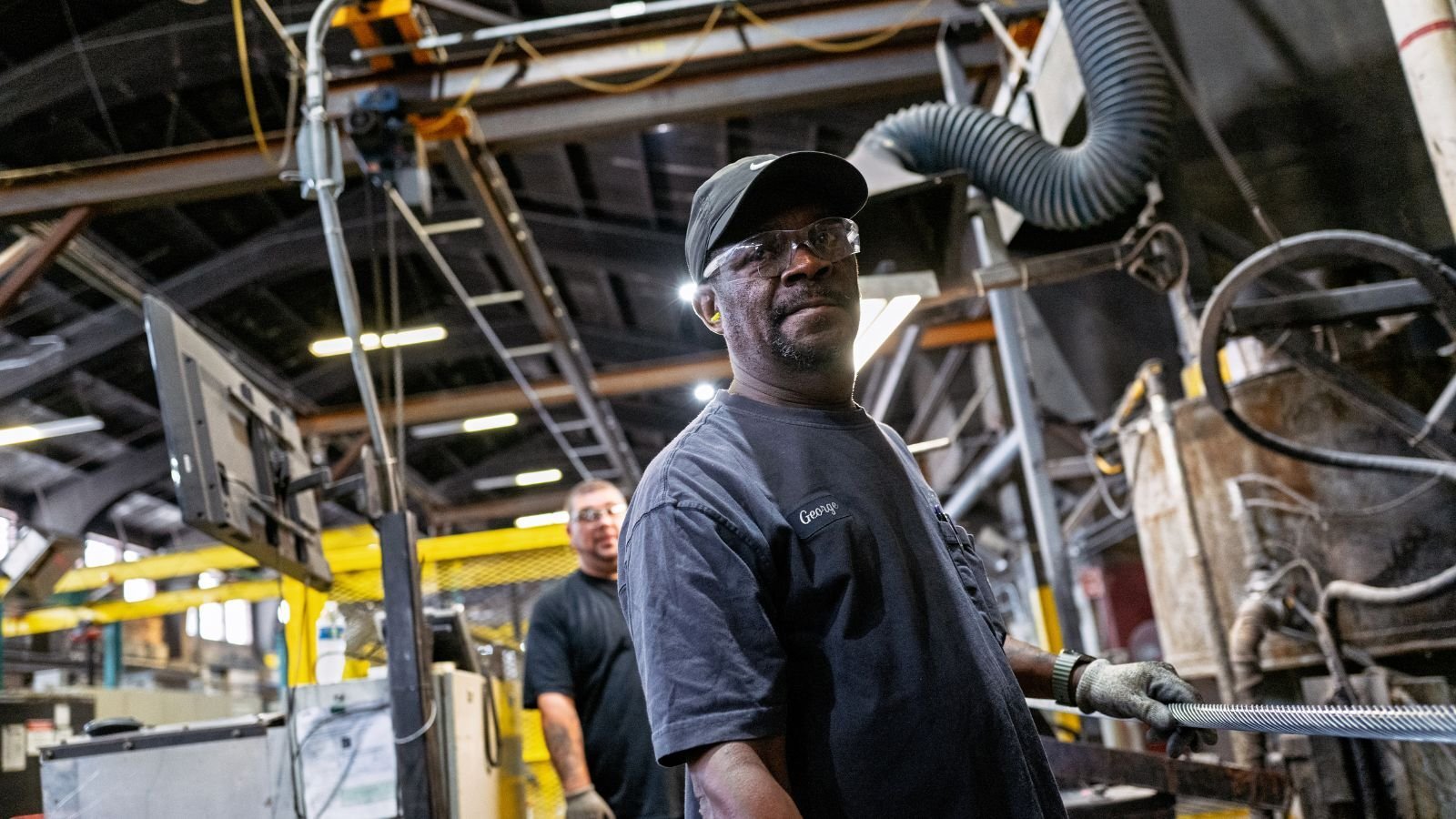 Two factory workers stand in an industrial setting with machinery and equipment. One man looks towards the camera.