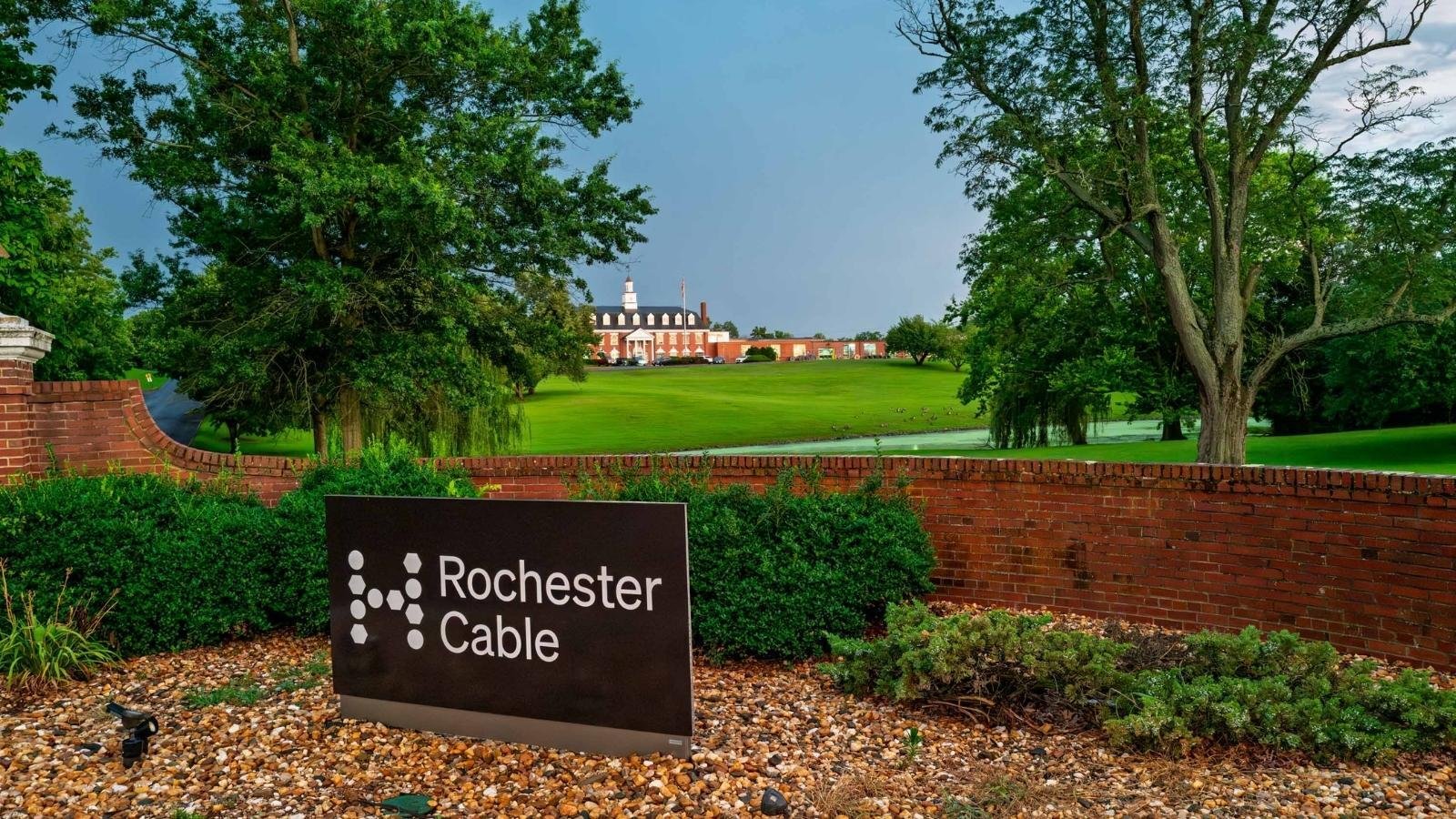A lush green landscape with trees frames a historic brick building in the distance. In the foreground, a sign reads 