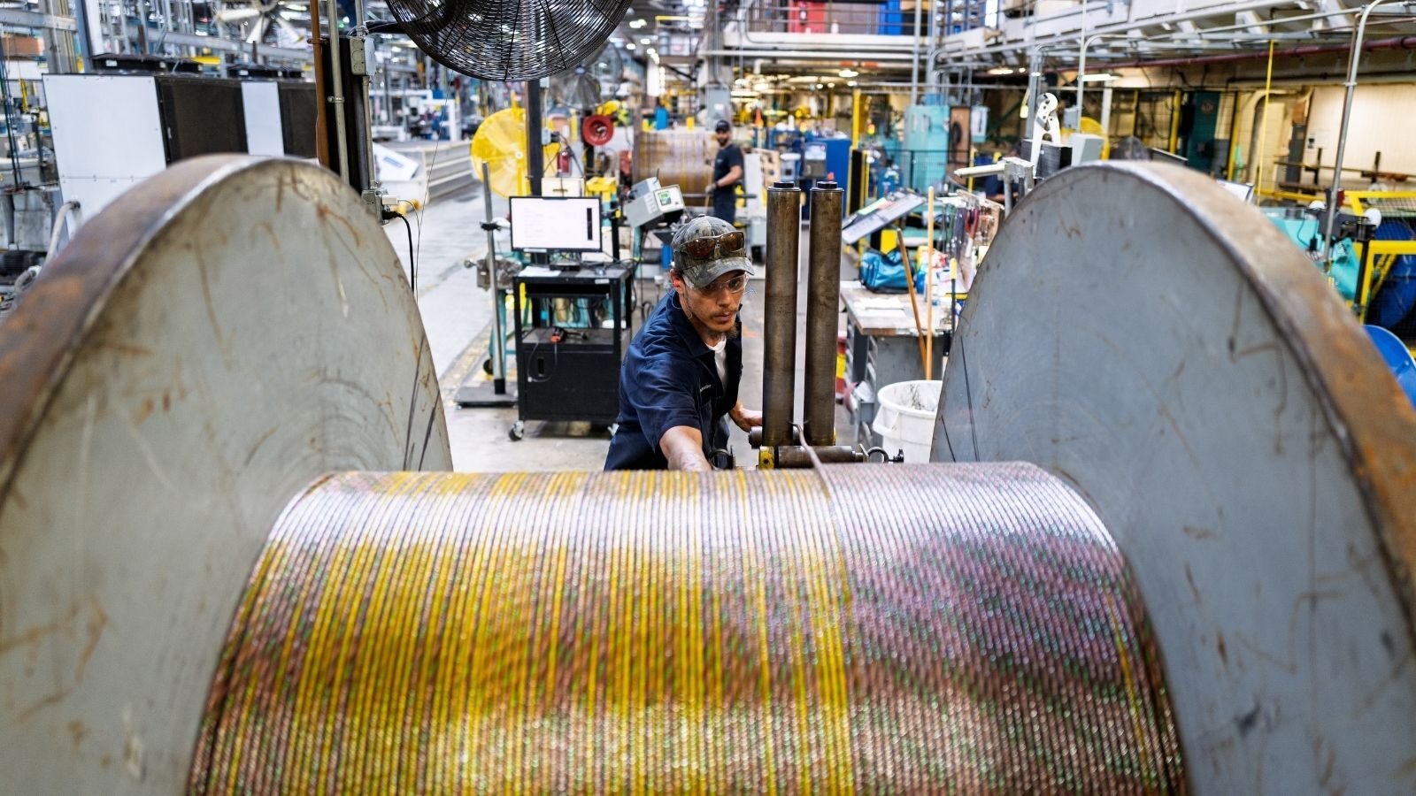 Factory worker operating machinery in an industrial setting. The focus is on large spools of colorful cables. The atmosphere is busy and industrious.
