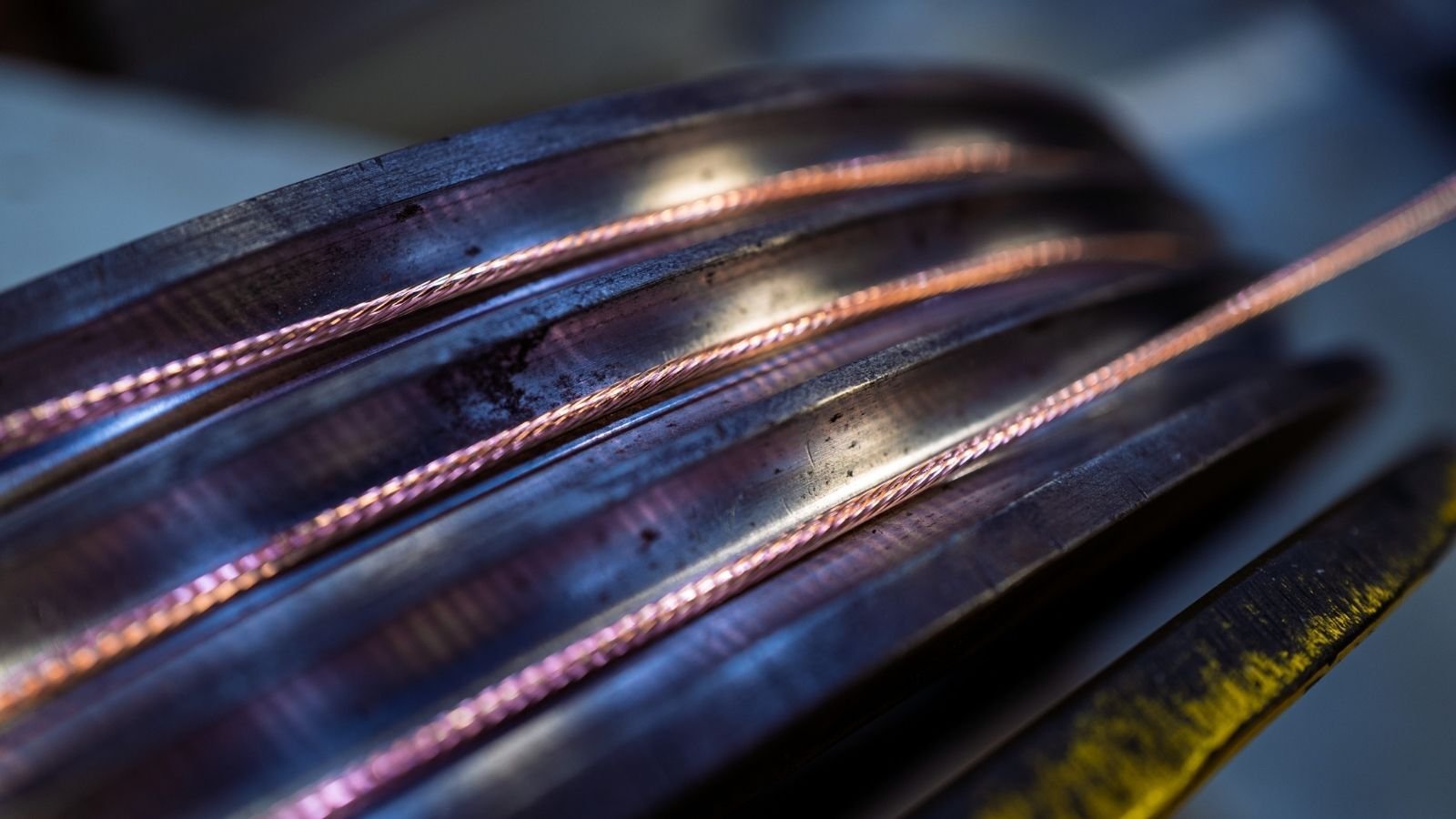 Close-up of coiled copper wires with a metallic sheen, set against a blurred industrial background. The lighting highlights their smooth, reflective surface.