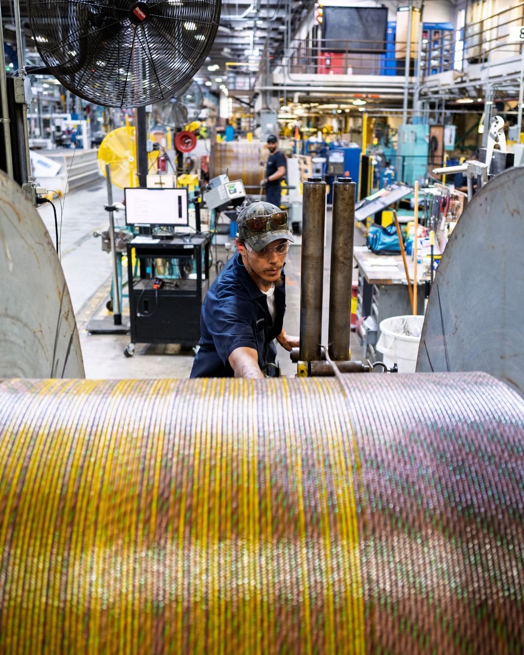 A factory worker operates machinery with large spools of multicolored cable in an industrial setting. The scene is busy and focused, with tools and equipment surrounding him.
