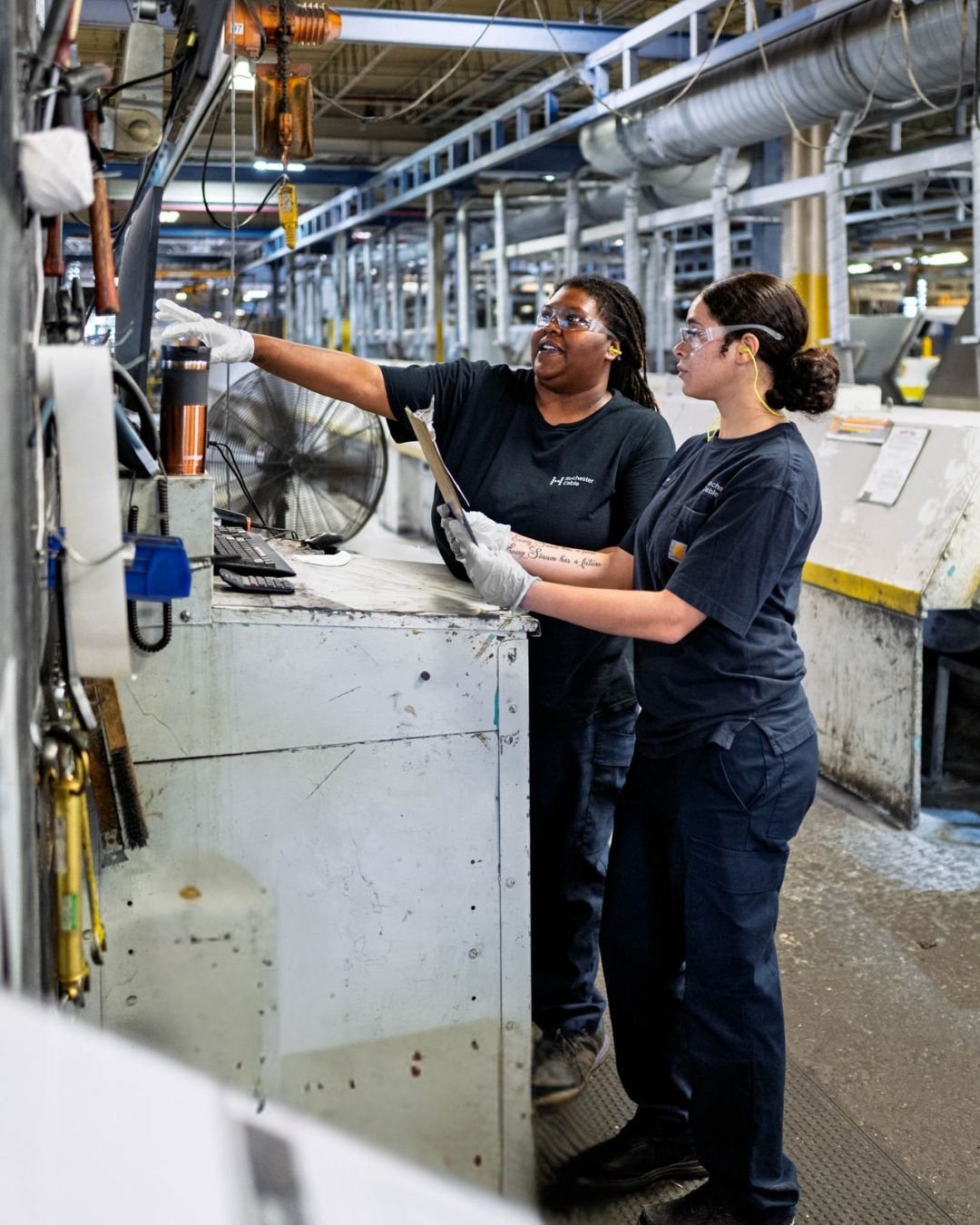 Two factory workers, wearing safety glasses and gloves, engage in discussion at a workstation.