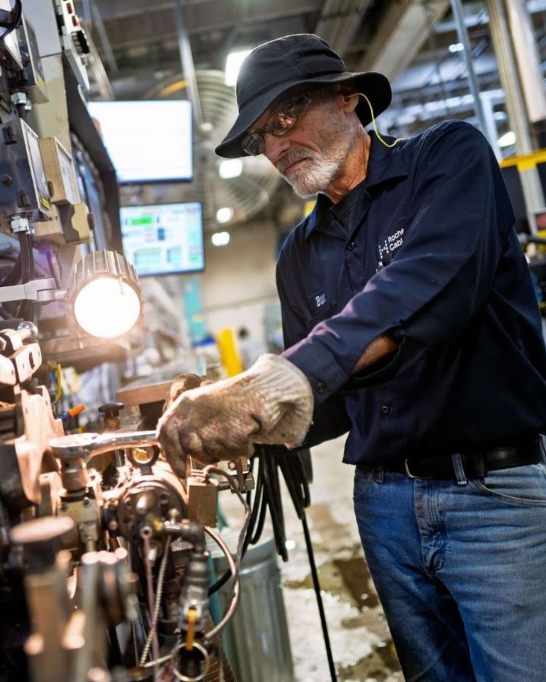 A man in a dark hat and safety glasses operates machinery in a factory. He wears gloves, focusing intently, with screens and industrial equipment around him.