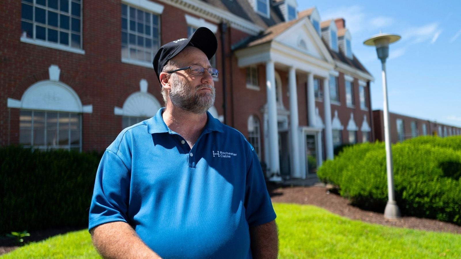 A man in a blue polo and cap stands in front of a large brick building with white columns, under a clear blue sky.