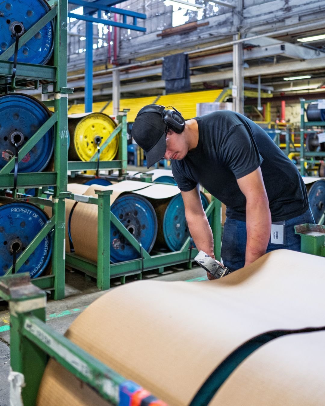 A worker in a black cap and ear protection inspects large paper rolls at a factory. Metal shelves with vibrant cable spools are in the background.