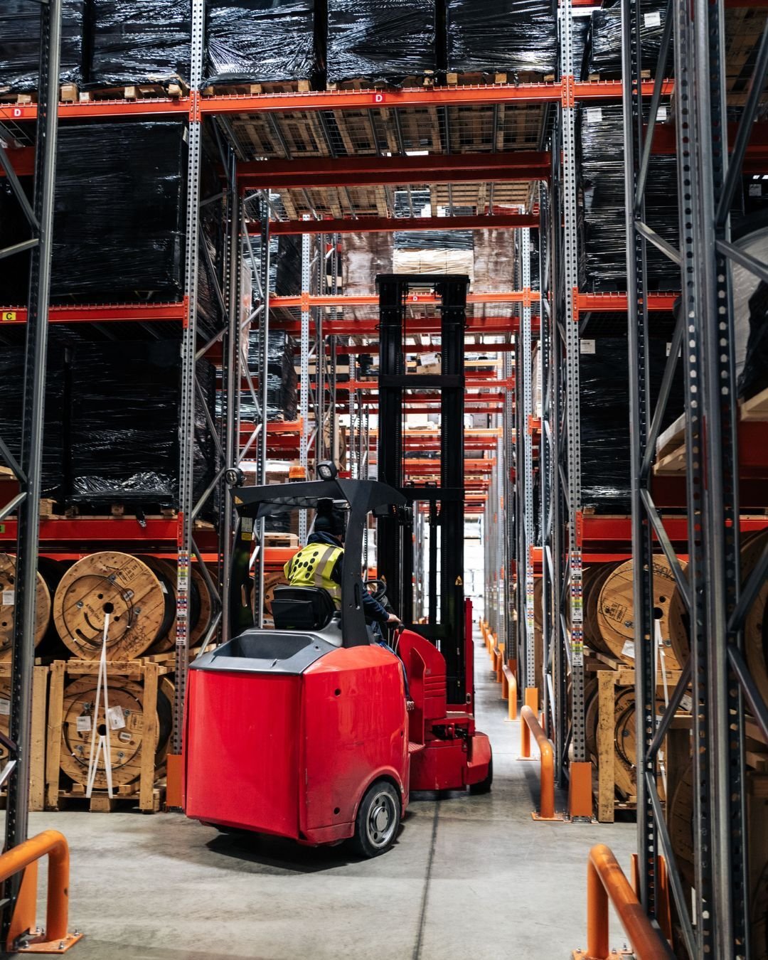 A worker operates a red forklift in a warehouse aisle, surrounded by tall metal shelves stacked with black-wrapped pallets and large wooden spools.