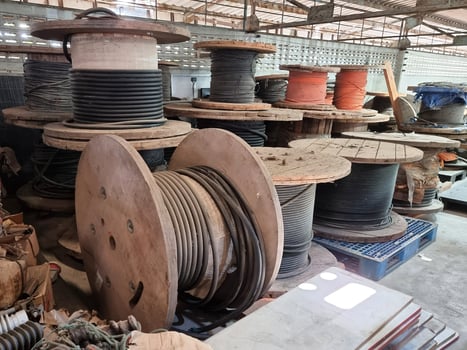 Many wooden cable spools filled with various colored electrical cables are stacked in a warehouse.