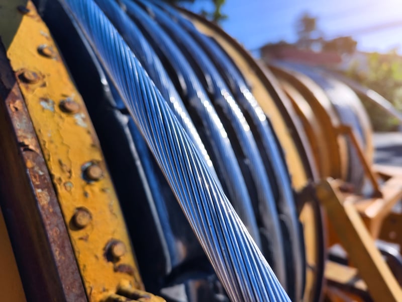 Close-up of thick, shiny steel cables wound around a metal spool, with an industrial yellow frame and sunlight reflecting off the surface.